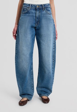Kvinde iført højtaljede, løstsiddende blå jeans med forlommer og brune flade sko, stående mod en ensfarvet baggrund.