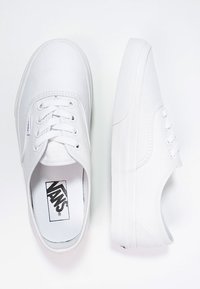 White canvas sneakers with a low profile, round toe, classic lace-up design, and textured rubber sole. Branding visible on the insole.