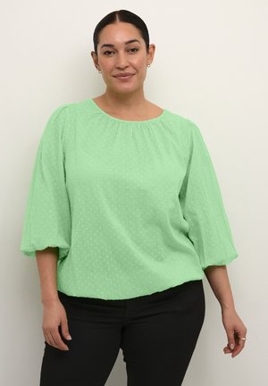 Chemisier vert menthe avec des pois texturés, col rond et manches bouffantes trois-quarts ; associé à un pantalon noir. Coupe décontractée.