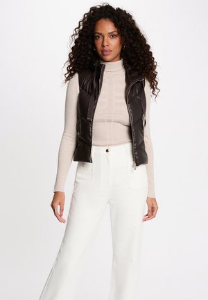Donna con capelli ricci che indossa un dolcevita beige, un gilet imbottito nero senza maniche e pantaloni bianchi a vita alta, in piedi davanti a uno sfondo semplice.