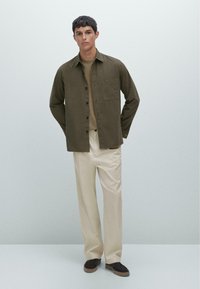 Khaki overhemd met knopen, beige ronde halstrui, crèmekleurige broek en donkerbruine sneakers. Het overhemd heeft een enkele borstzak en een ontspannen pasvorm.