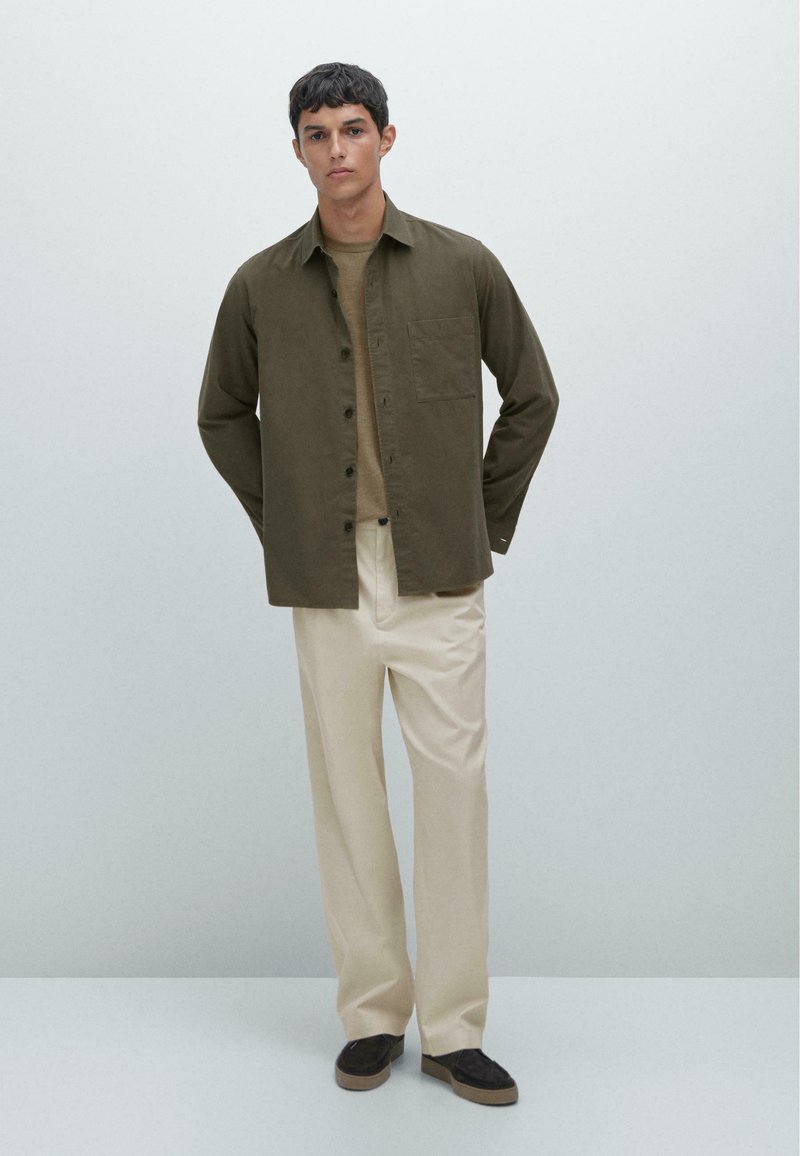 Khaki overhemd met knopen, beige ronde halstrui, crèmekleurige broek en donkerbruine sneakers. Het overhemd heeft een enkele borstzak en een ontspannen pasvorm.