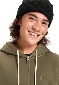 Sudadera con cremallera en verde oliva con cordones en color crema y logo de Quiksilver, llevada con un gorro de punto negro. Presenta una textura de tela suave.