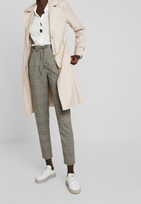 Beige trenchcoat över en vit blus med knappdetaljer, tillsammans med grå rutiga högmidjade byxor och vita sneakers.