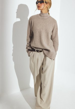 Ljusbrun stickad tröja med ribbat nederkant och hög krage, ihop med lösa beiga byxor och ett mörkt bälte, mot en vit bakgrund.