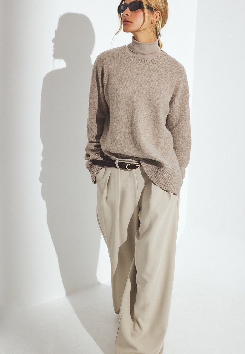Heled pruun kudumissweater ribatud ääre ja kõrge kaelusega, kombineeritud laia beeži püksiga ja tumeda vööga, valge taustaga.