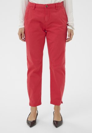 Moteris, dėvinti ryškiai raudonas tiesias kelnes su šoninėmis kišenėmis, derinamas su juodais bateliais su smailiomis nosytėmis, stovinti priešais paprastą foną.