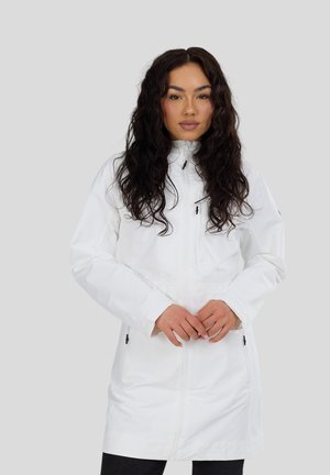 Kobieta o długich, kręconych ciemnych włosach, ubrana w białą kurtkę na zamek, stojąca na tle jednolitego jasnoszarego tła.