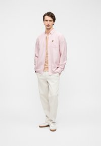 Ανοιχτό ροζ πουκάμισο με λεπτές ρίγες, τσέπη και μικρό μαύρο λογότυπο, φορεμένο πάνω από μπεζ μπλουζάκι και σε συνδυασμό με ανοιχτόχρωμο παντελόνι.