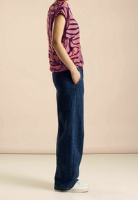 Pink top with dark wavy pattern, short sleeves; paired with loose blue jeans and white sneakers. Side view against a neutral background.