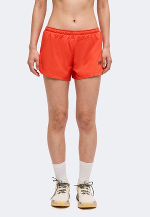 Personne portant un short de sport orange vif, des chaussettes blanches et des chaussures de course de couleur claire, debout devant un fond uni.