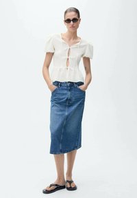 Blusa bianca con texture, maniche corte a sbuffo e dettaglio a chiave sul davanti, abbinata a una gonna di jeans blu fino al ginocchio e sandali neri.
