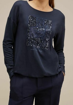Femme portant une chemise à manches longues bleu marine avec un motif carré de sequins et les mots "sourire · amour · plaisir" sur le devant, les mains dans les poches.