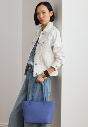 Femme portant une veste blanche, un haut bleu à fleurs et un jean, tenant un sac à main violet, debout contre un mur blanc à l'intérieur.