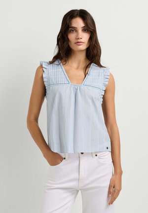 Vrouw met donker golvend haar, die een mouwloze lichtblauwe top met ruches en een witte broek draagt tegen een effen achtergrond.