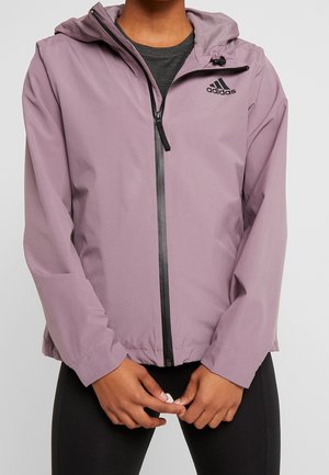Persoon draagt een mauve Adidas-hoodie met een zwarte ritssluiting, gecombineerd met een zwarte broek, met de handen voor zich samengevouwen.