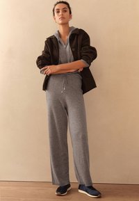 Gray knit jumpsuit with a hood and drawstring waist paired with an oversized brown jacket. Dark sneakers complete the outfit.