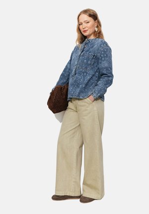 Femme portant une chemise en jean à motifs bleus, un pantalon beige à jambes larges, tenant un sac marron à imprimé léopard, debout avec une main dans la poche.
