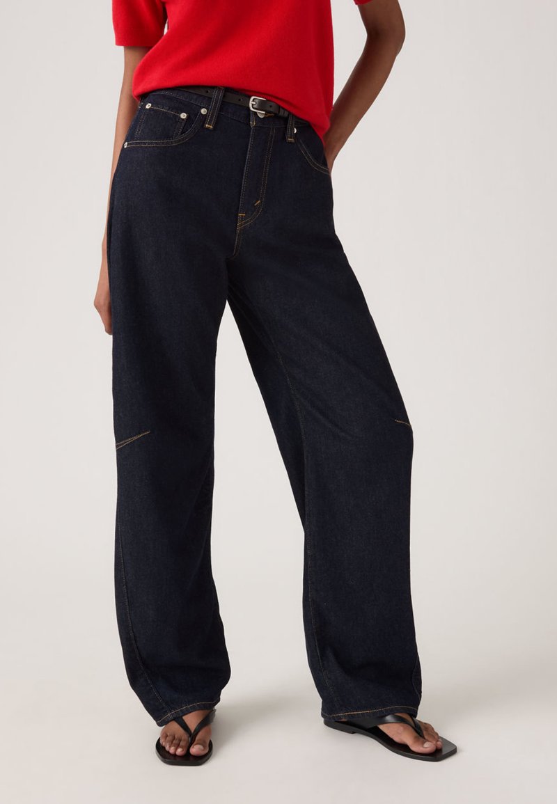 Τζιν παντελόνι με φαρδιά μπατζάκια από σκουρόχρωμο τζιν με εμφανείς ραφές, σχέδιο με πέντε τσέπες και ψηλή μέση. Συνδυασμένο με κόκκινη μπλούζα και μαύρες σαγιονάρες.