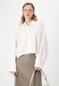 Weißes, kurzes Button-Up-Hemd mit langen Ärmeln, mit einem klassischen Kragen und subtilen Glanz, kombiniert mit einem beigen Rock.