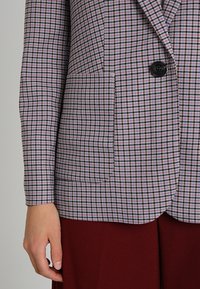 Fialový a černý kostičkovaný blazer se zapínáním na jeden knoflík, dvěma předními kapsami a hladkým povrchem. V kombinaci s vínovými kalhotami.