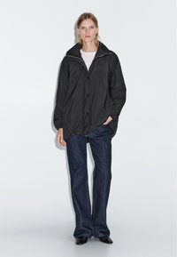 Una donna sta in piedi indossando una giacca nera oversize sopra una camicia bianca, jeans dritti blu scuro e scarpe nere a punta su uno sfondo semplice.