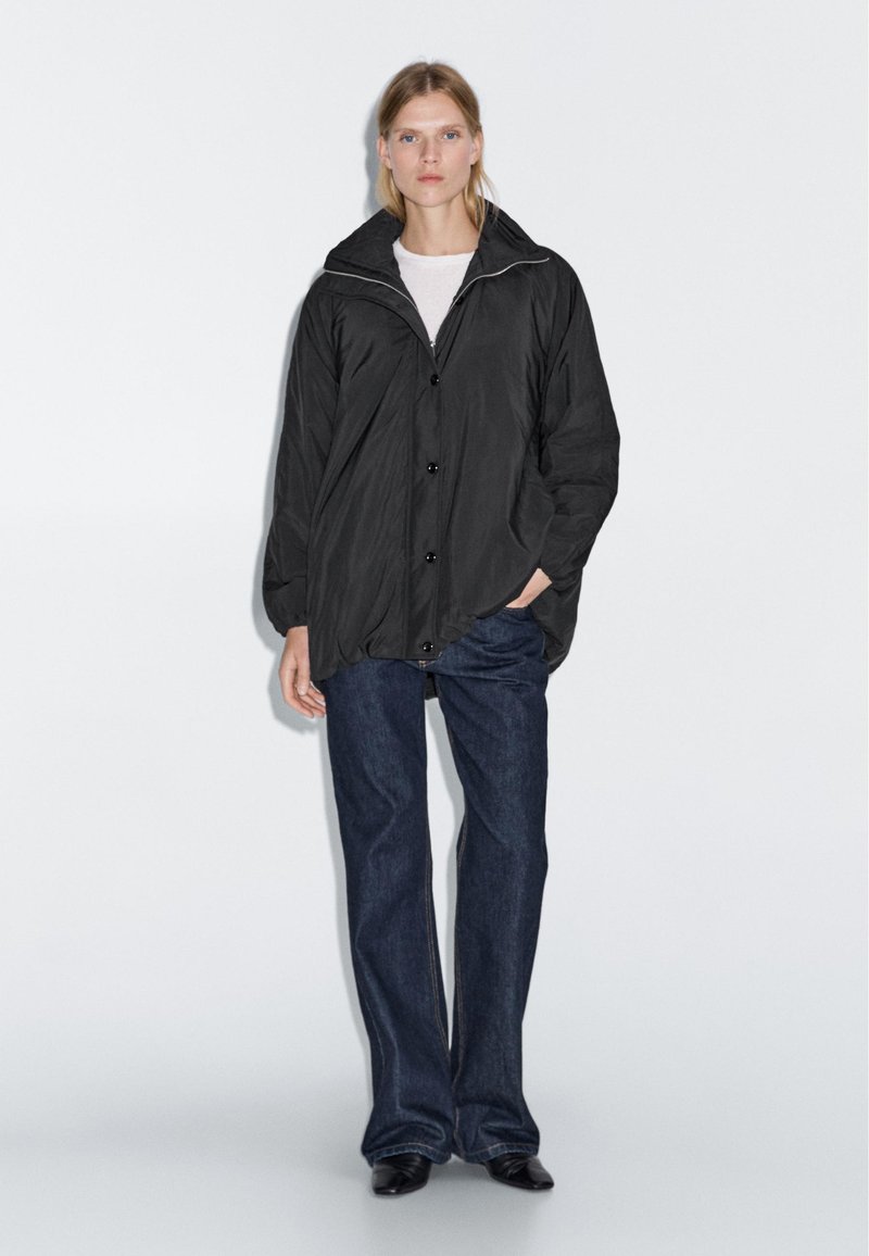 Kvinden står iført en sort oversized jakke over en hvid skjorte, mørkeblå lige ben jeans og sorte spidse sko foran en ensfarvet baggrund.