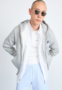 Siva zip-up kapucar preko bele prugaste košulje i bele majice bez rukava, u kombinaciji sa svijetloplavim trenerkama i okruglim naočarima.