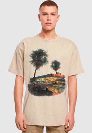 Beige bomulds t-shirt med et grafik af palmer, en retro bil og en butik med teksten "2Y STUDIOS," sat mod en regnfuld scene.