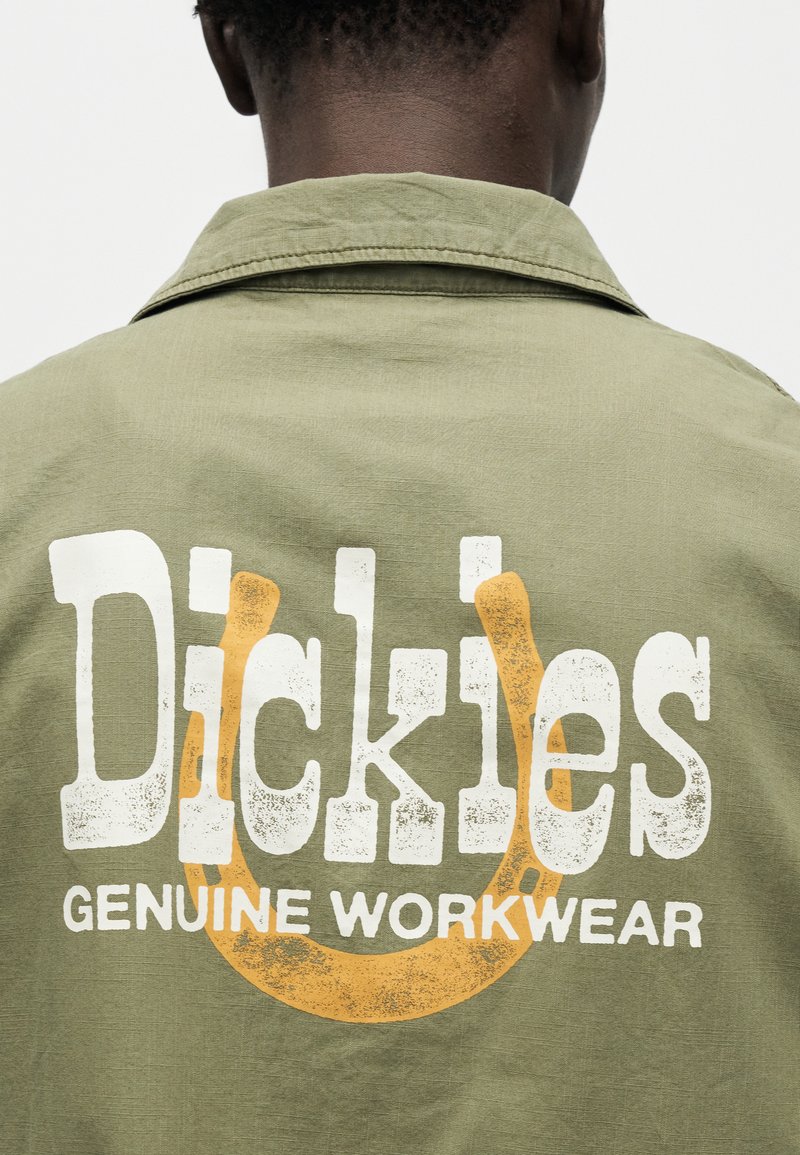 Vista trasera de una persona que lleva una chaqueta verde oliva con el texto "Dickies Genuine Workwear" y un gran logo naranja de herradura en la parte posterior.