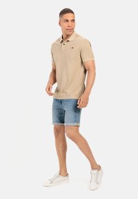 Beige poloshirt met een kraag en drie knopen, gecombineerd met lichtblauwe denimshorts. Accenten zijn een klein logo en oprolbare zoom. Witte sneakers.