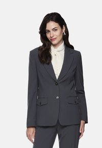 Gray suit jacket with a notched lapel, single button closure, and two front pockets. Worn over a white blouse with a tied neck accent.