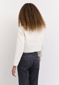 Gekürzte, strukturierte Jacke aus off-weißem Stoff mit langen Ärmeln, kombiniert mit hochgeschnittenen, dunklen Jeans. Einfaches Design ohne sichtbare Verschlüsse.