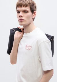 Λευκό βαμβακερό μπλουζάκι με κόκκινο κείμενο. Το μοντέλο κρατάει μια μαύρη τσάντα με λεία υφή και φερμουάρ, παρουσιάζοντας έναν μινιμαλιστικό σχεδιασμό.