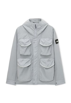 Lichtgrijze jack met capuchon en vier grote klepzakken aan de voorkant, en een zwart label op de linkermouw, voorzien van een ritssluiting en drukknoopsluiting.