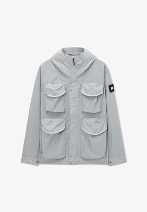 Vaaleanharmaa huppari, jossa on neljä suurta etutaskua läpällä ja musta merkki vasemmassa hihassa, vetoketju- ja napillissuljenta.