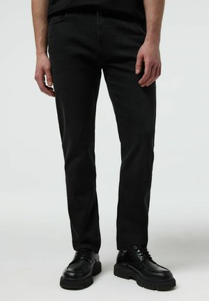 Mężczyzna noszący czarne jeansy o prostym kroju i czarne skórzane sznurowane buty, stojący na jednolitym jasnym tle.