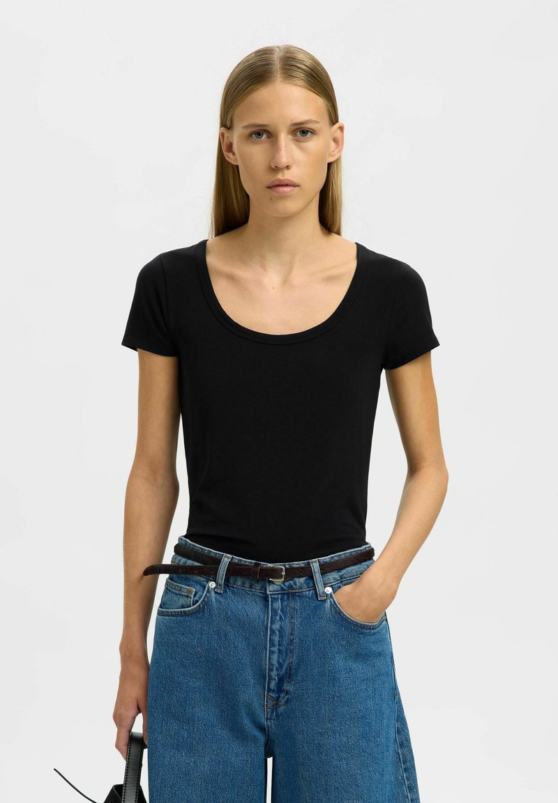 Sort t-shirt med korte ærmer og dyb rund halsudskæring, parret med højtaljede blå denimjeans, der har en afslappet pasform og diskret tekstur.