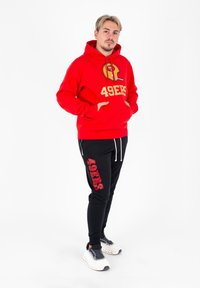 Czerwona bluza z kapturem z złotym logo 49ERS i grafiką hełmu, zestawiona z czarnymi spodniami dresowymi z czerwonym napisem 49ERS. Obuwie to białe buty sportowe.