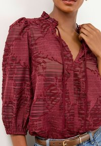 Gros plan d'une personne portant un haut transparent texturé bordeaux à manches 3/4, avec un col volan, un décolleté à cordon, associé à une ceinture et un jean.
