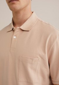 Beige színű pólóing texturált anyagból, gallérral, három gombbal és egy bal oldali melletti zsebbel, finom logóval.