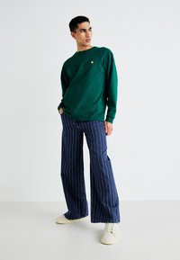 Green long-sleeve shirt with a small logo at the chest, paired with wide-leg navy striped jeans and cream-colored sneakers.