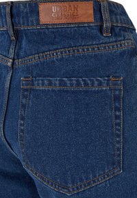Poche arrière et taille avec étiquette en cuir marron portant la mention Urban Classics Denim Factory sur un jean en denim bleu avec des surpiqûres orange.