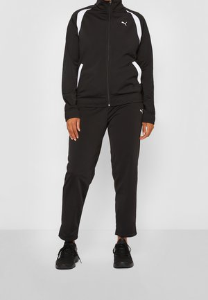 Personne portant un survêtement noir Puma avec des accents blancs aux épaules et aux poches, associée à des chaussures de sport noires, debout devant un fond uni.