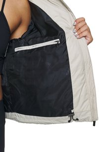 Leichte beige Jacke mit glattem schwarzen Innenfutter. Verfügt über eine Reißverschlusstasche und einen verstellbaren Kordelzug am Saum.