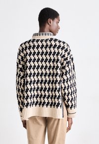 Strickpullover mit einem beigen und schwarzen Kreuzmuster, ausgestattet mit einem gerippten Saum und Kragen, entworfen mit einem lockeren Schnitt und seitlichen Schlitzen.