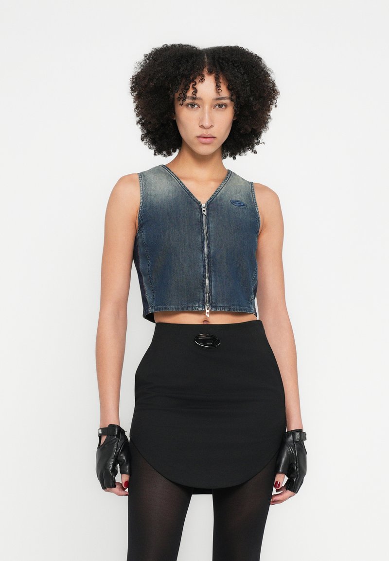 Kék farmer rövid mellény cipzáros elülső résszel, V-nyakú, fekete szoknyával párosítva, amely íves szegéllyel és kerek fekete fém díszítéssel rendelkezik.