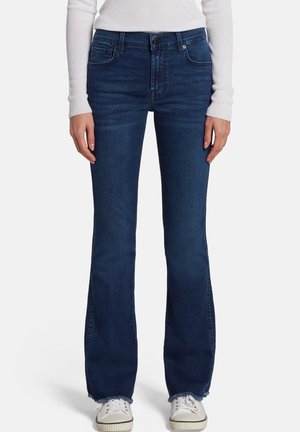 Femme portant un jean bootcut bleu foncé, un haut blanc à manches longues côtelé et des baskets blanches, debout avec les mains détendues le long du corps.