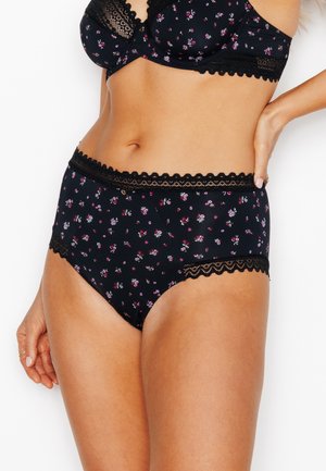 Femme portant un ensemble de lingerie en dentelle noire à motifs floraux, main sur la hanche, montrant la partie médiane sur un fond blanc uni.