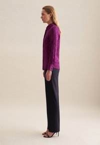 Blusa a fantasia fucsia con colletto alto, abbinata a pantaloni neri dritti e sandali neri con tacco, mostrata di lato.